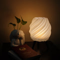 A glowing spiral ribbed table lamp with a white shade and black tripod base sits on a dark table next to a small white ceramic plant holder with green leaves and a red book.